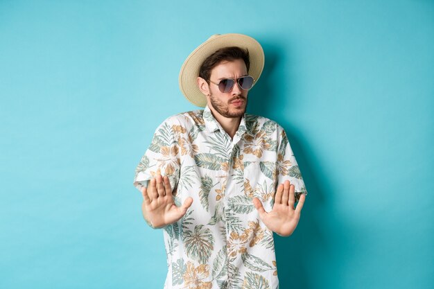 Turista alarmado que pide mantenerse alejado, un paso atrás de algo encogerse, mostrando un gesto de rechazo, de pie con sombrero de paja y camisa hawaiana, fondo azul.