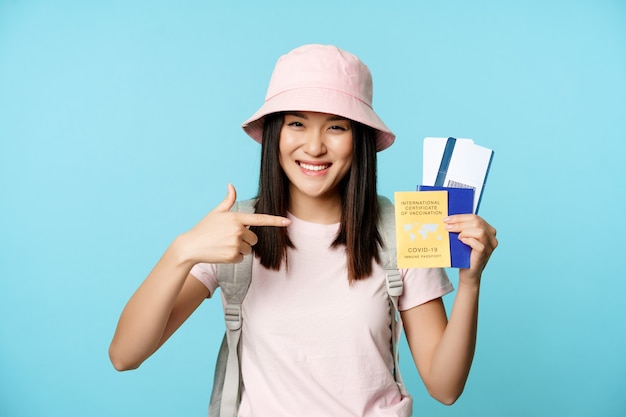 Turista asiático vacunado, niña mostrando pasaporte sanitario internacional, boletos para gira en el extranjero, sonriendo feliz, viajando durante la pandemia del covid-19, fondo azul.