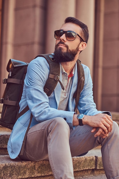 Foto gratuita un turista con barba y corte de pelo, vestido con ropa informal con mochila y gafas de sol, sentado en un escalón en una calle antigua.