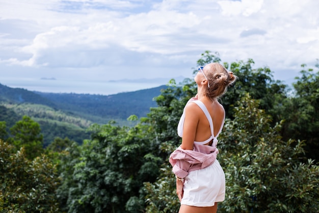 Turista de blogger de mujer con estilo europeo se encuentra en la cima de la montaña con una increíble vista tropical de la isla de Koh Samui Tailandia Moda retrato al aire libre de mujer