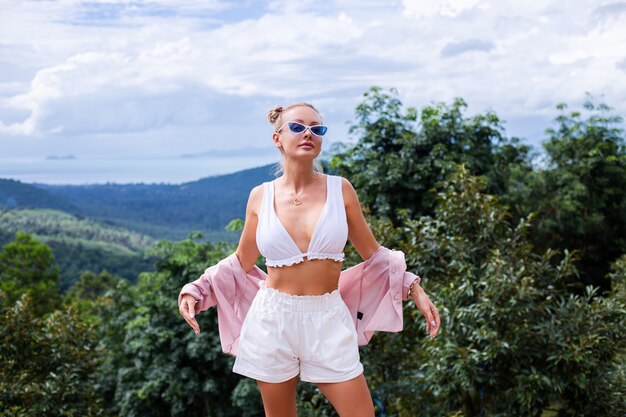 Turista de blogger de mujer con estilo europeo se encuentra en la cima de la montaña con una increíble vista tropical de la isla de Koh Samui Tailandia Moda retrato al aire libre de mujer