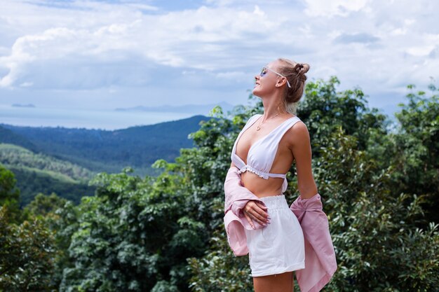 Turista de blogger de mujer con estilo europeo se encuentra en la cima de la montaña con una increíble vista tropical de la isla de Koh Samui Tailandia Moda retrato al aire libre de mujer