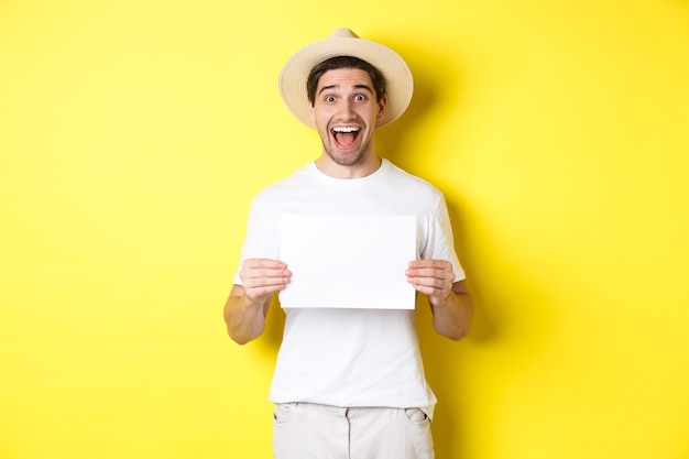 Turista emocionado mostrando su logotipo o firmar en un trozo de papel en blanco, sonriendo asombrado, de pie contra el fondo amarillo.