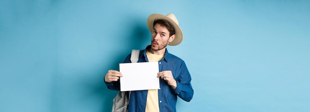 Turista emocionado con sombrero de paja haciendo autostop mostrando un trozo de papel en blanco y luciendo divertido de pie