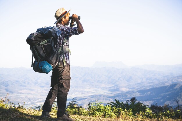 El turista está mirando a través de los prismáticos en el cielo nublado soleado desde la cima de la montaña.