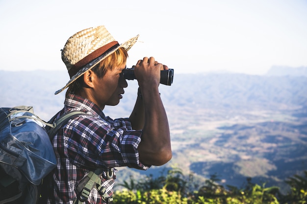 El turista está mirando a través de los prismáticos en el cielo nublado soleado desde la cima de la montaña.