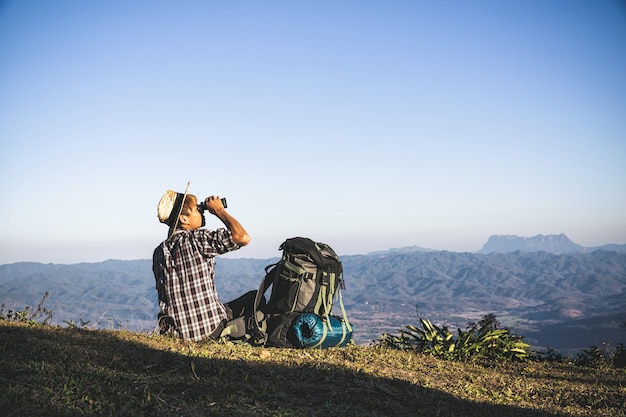 El turista está mirando a través de los prismáticos en el cielo nublado soleado desde la cima de la montaña.