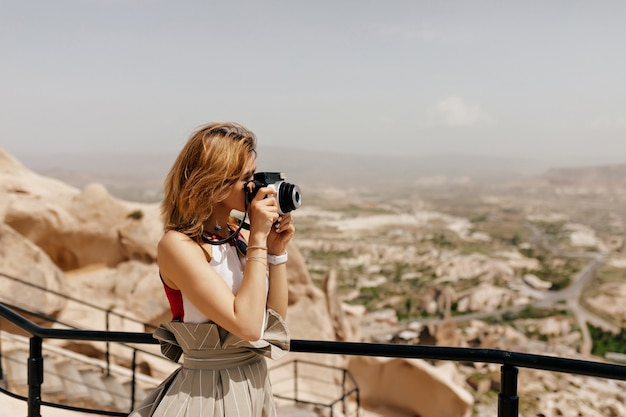 Turista con estilo de pelo corto está haciendo fotos entre rocas antiguas con vistas a la ciudad a la luz del sol