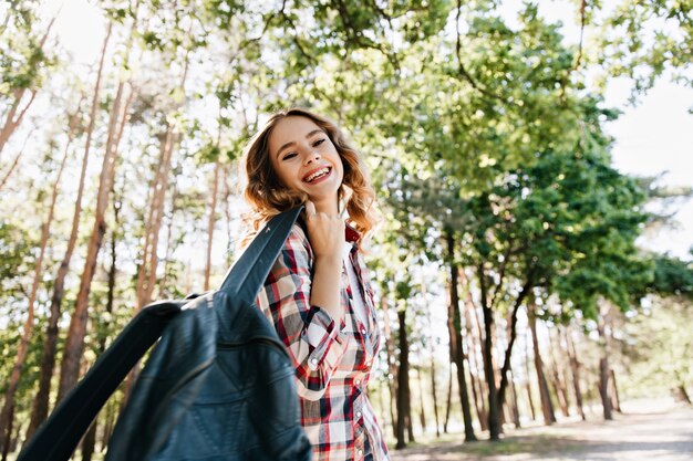 Turista Jocund divirtiéndose en la naturaleza. Foto al aire libre de la hermosa chica rubia con mochila riendo en un día soleado.