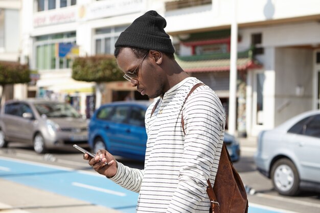 Turista joven negro de moda con mochila de cuero mirando el teléfono inteligente en sus manos con expresión seria, utilizando la aplicación de navegación en línea, buscando dirección mientras se pierde en la gran ciudad