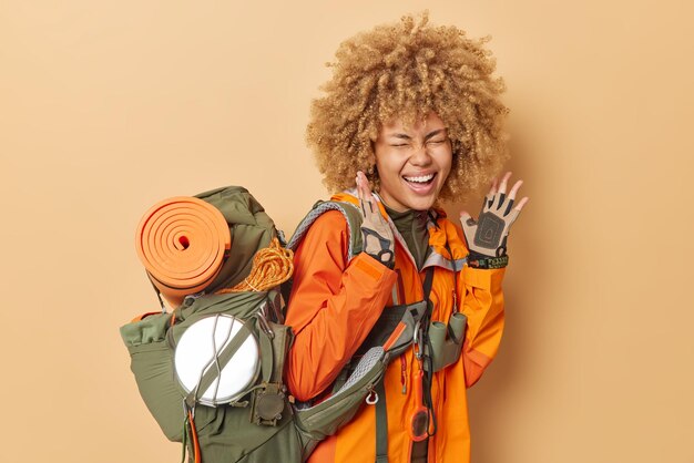 Una turista llena de alegría exclama en voz alta mantiene las manos levantadas usa guantes chaqueta naranja lleva una mochila pesada con el equipo necesario aislado sobre fondo marrón Concepto de aventura de verano