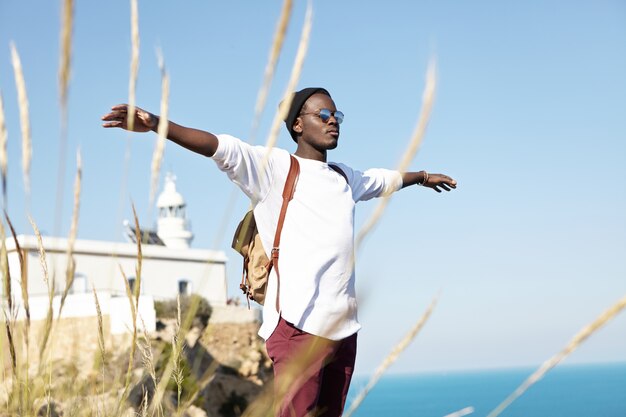 Turista masculino elegante feliz y libre que tiene una mirada relajada y despreocupada mientras está parado en el borde del acantilado, extendiendo los brazos como un pájaro, sintiendo el viento cálido en un día soleado durante su viaje al extranjero. Concepto de verano