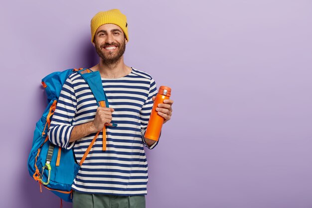 Foto gratuita el turista masculino encantado y sonriente tiene un viaje increíble, lleva una mochila grande, bebe café del matraz, está de buen humor, usa un sombrero amarillo