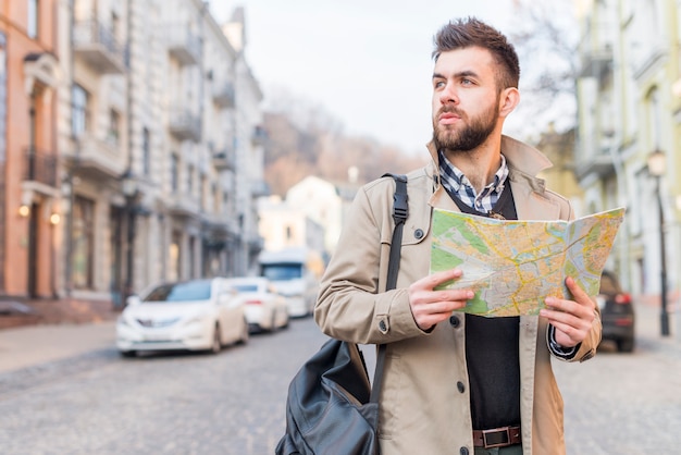 Turista masculino joven con el bolso en su hombro que se coloca en la calle que sostiene el mapa en la mano que mira lejos