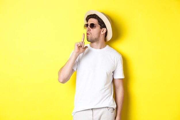 Turista masculino joven fresco que sopla en la pistola del dedo y que parece seguro, de pie contra el fondo amarillo. Concepto de vacaciones y estilo de vida