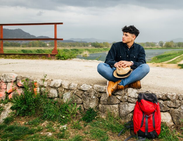 Turista masculino joven que se sienta con la mochila cerca del río hermoso