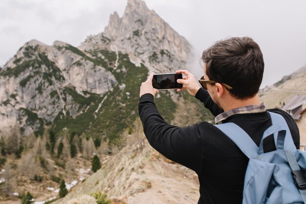 Turista masculino con pelo corto negro admira las montañas italianas y toma fotos en el teléfono inteligente
