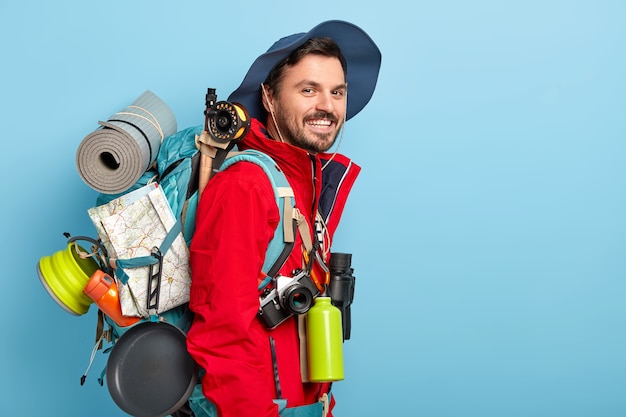 Foto gratuita turista masculino sonriente lleva sombrero y chaqueta roja, lleva mochila con mapa, karemat, utiliza binoculares, frasco con bebida caliente