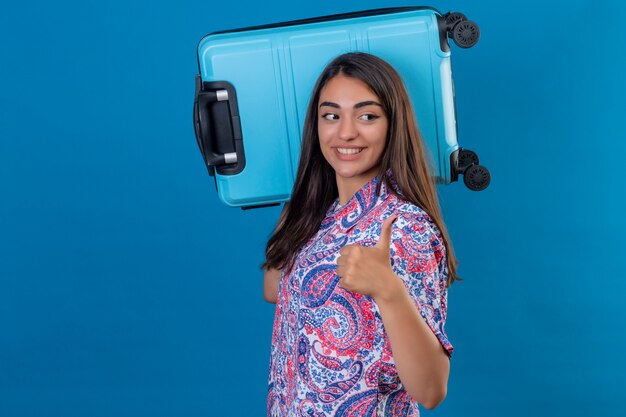 Foto gratuita turista mujer de pie con maleta de viaje en el hombro sonriendo alegremente mostrando thumbs up mirando a un lado en azul aislado