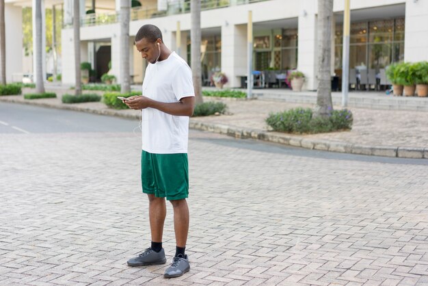 Turista negro enfocado preparándose para trotar y establecer la lista de reproducción en el teléfono.