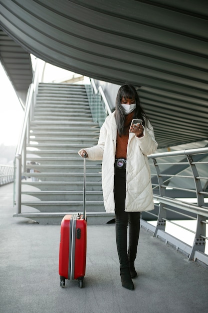 Foto gratuita turista revisando el teléfono inteligente y sosteniendo el equipaje mientras usa una máscara médica