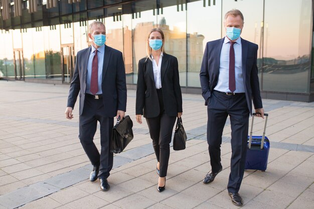 Turistas de negocios con máscaras faciales que visitan la oficina de socios extranjeros, maletas con ruedas, caminar al aire libre. Vista frontal. Viaje de negocios y concepto de epidemia.