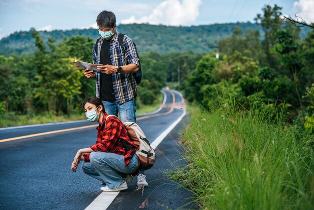 Turistas sentados, turistas masculinos mirando el mapa, ambos con máscaras y al costado de la carretera.