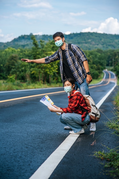 Las turistas se sientan y miran el mapa, los turistas masculinos fingen hacer autostop. Ambos usan máscaras y están al costado del camino.