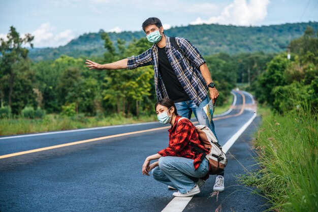 Las turistas se sientan, los turistas masculinos, fingen ser autostopistas, ambos con una máscara y al costado de la carretera.