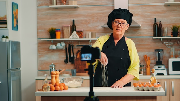 Tutorial sobre harina mientras se registra la preparación de alimentos en la cocina de casa. Influencer chef blogger jubilado que usa la tecnología de Internet para comunicarse, grabar blogs en las redes sociales con equipos digitales