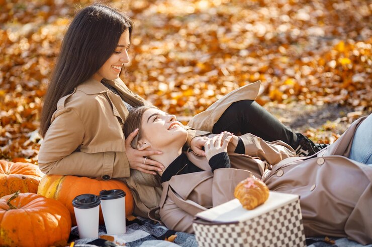 Photo Gratis two beautiful female friends spending time on a picnic blanket on the grass. two young smiling sisters making picnic and hugging in autumn park. brunette and blonde girls wearing coats.