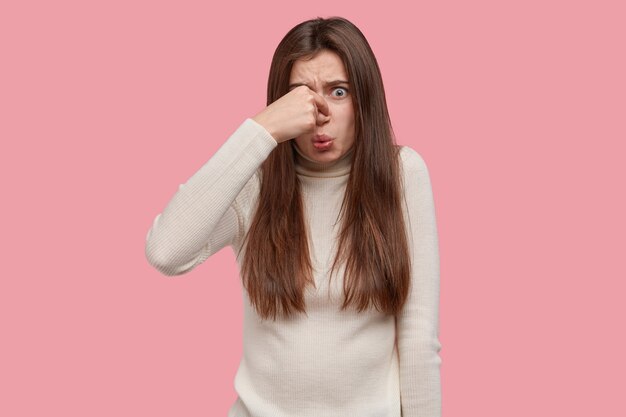 Uf, qué olor repugnante. Descontento mujer morena cubre la nariz, siente mal hedor, viste un suéter blanco informal de cuello alto, nota productos podridos