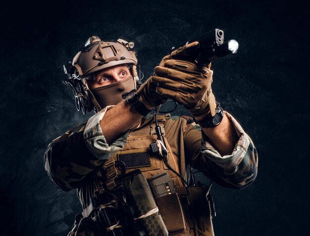 Unidad de élite, soldado de las fuerzas especiales con uniforme de camuflaje sosteniendo un arma con una linterna y apuntando al objetivo. Foto de estudio contra una pared de textura oscura