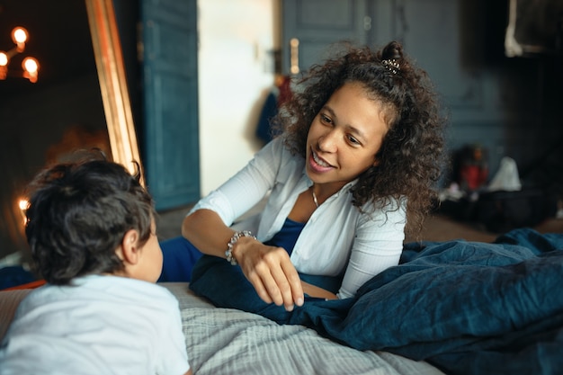 La unión entre madre e hijo en el dormitorio. Feliz cuidado joven mujer de raza mixta relajante en casa con su pequeño hijo