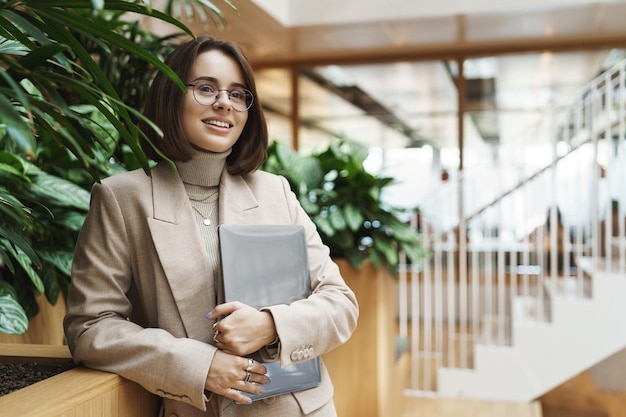 Foto gratuita universidad de carrera y concepto de estilo de vida una joven inteligente soñadora y optimista logra el objetivo de postularse para un puesto de trabajo de pie en la recepción cerca de las plantas con una computadora portátil en las manos sonriendo feliz