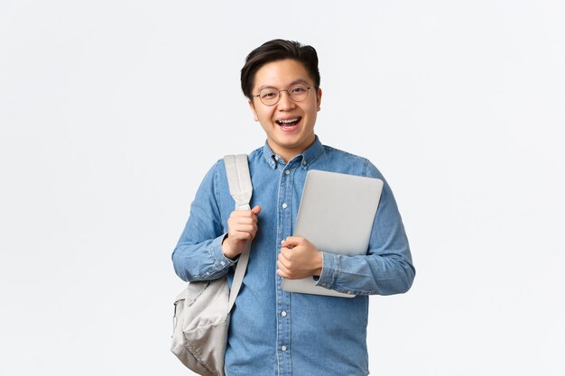 Universidad, estudio en el extranjero y concepto de estilo de vida. Chico asiático alegre sonriente en vasos de pie con mochila y portátil. Estudiante en su camino a clases, posando sobre fondo blanco.