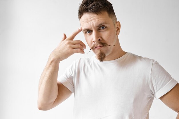 Usa tu cerebro. Retrato de estudio de hombre guapo emocional con barba recortada y bigote de manillar con mirada disgustada, sosteniendo el dedo índice en su sien y rodándolo, diciendo: ¿Estás loco?