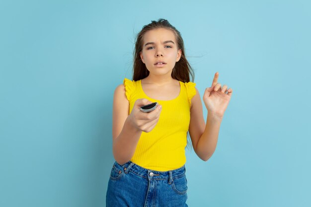 Usando el control remoto de la TV. Retrato de muchacha adolescente caucásica aislado en azul