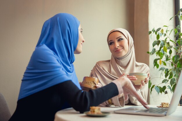 Usando dispositivos. Hermosas mujeres árabes reunidas en una cafetería o restaurante, amigos o reuniones de negocios. Pasar tiempo juntos, hablando, riendo. Estilo de vida musulmán. Modelos elegantes y felices con maquillaje.