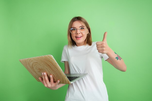 Usando laptop emocional. Retrato de mujer joven caucásica aislado en la pared verde. Modelo de mujer hermosa en camisa blanca. Concepto de emociones humanas, expresión facial, juventud.