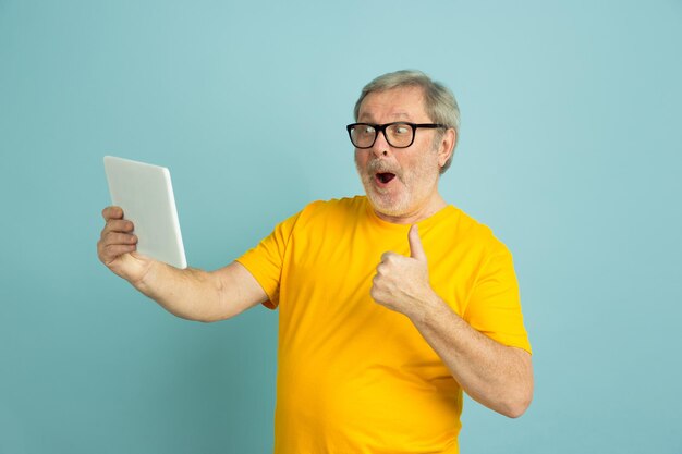 Usando tableta, mostrando el pulgar hacia arriba. Retrato de hombre caucásico aislado sobre fondo azul de estudio. Hermoso modelo masculino en camisa amarilla.