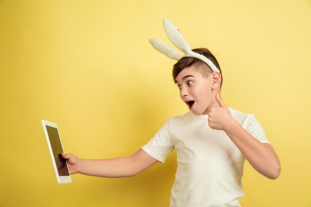 Usando tableta, pulgar hacia arriba. Chico caucásico como un conejito de Pascua sobre fondo amarillo de estudio. Saludos felices de pascua.