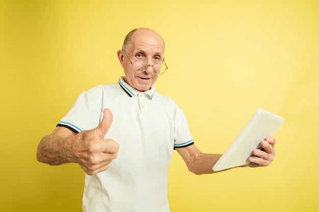 Usando tableta, pulgar hacia arriba. Retrato de hombre mayor caucásico sobre fondo amarillo de estudio. Hermoso modelo emocional masculino. Concepto de emociones humanas, expresión facial, ventas, bienestar, publicidad. Copyspace.