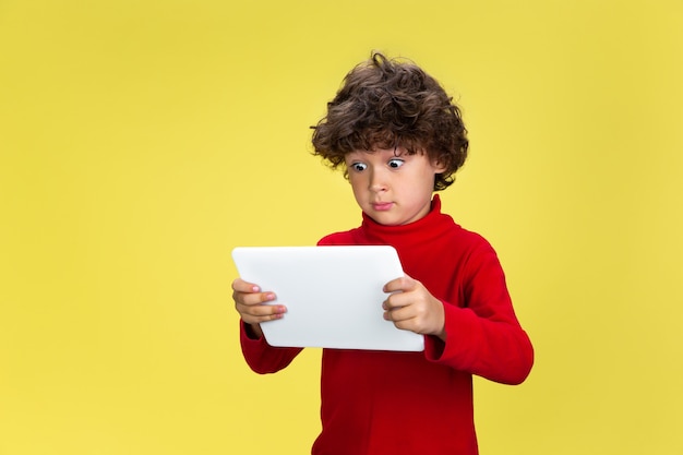 Foto gratuita usando tableta. retrato de niño rizado bastante joven en suéter rojo en la pared amarilla del estudio