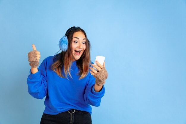 Usando el teléfono, feliz. Retrato de mujer caucásica sobre fondo azul de estudio. Modelo de mujer hermosa en ropa de abrigo. Concepto de emociones humanas, expresión facial, ventas, publicidad. Estado de ánimo de invierno, vacaciones.