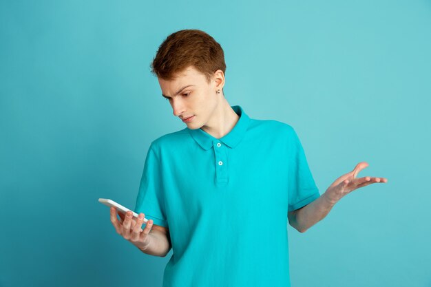 Usando el teléfono. Retrato moderno del hombre joven caucásico aislado en la pared azul, monocromo. Hermoso modelo masculino. Concepto de emociones humanas, expresión facial, ventas, publicidad, moda.