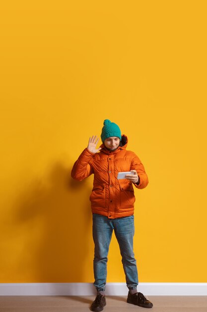 Usar gadgets en cualquier lugar. Hombre caucásico joven con smartphone, sirviendo, charlando, apostando. Retrato de cuerpo entero aislado en la pared amarilla. Concepto de tecnologías modernas, millennials, redes sociales.