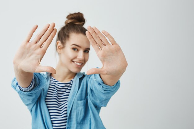 Usted es muy fotogénico. Foto de estudio de la bella y creativa fotógrafa con peinado de moño, tirando de las palmas hacia la cámara, midiendo el ángulo de disparo, sonriendo ampliamente y de buen humor