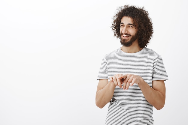 Ustedes dos durmieron juntos, ¿verdad? Retrato de intrigado alegre amigo masculino con corte de pelo rizado y barba, frotando el dedo índice y riendo con alguna pista