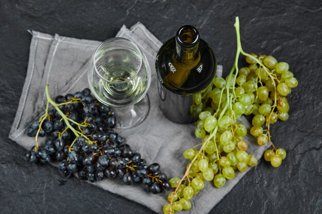 Uva mixta, una copa de vino y una botella con mantel gris sobre superficie oscura